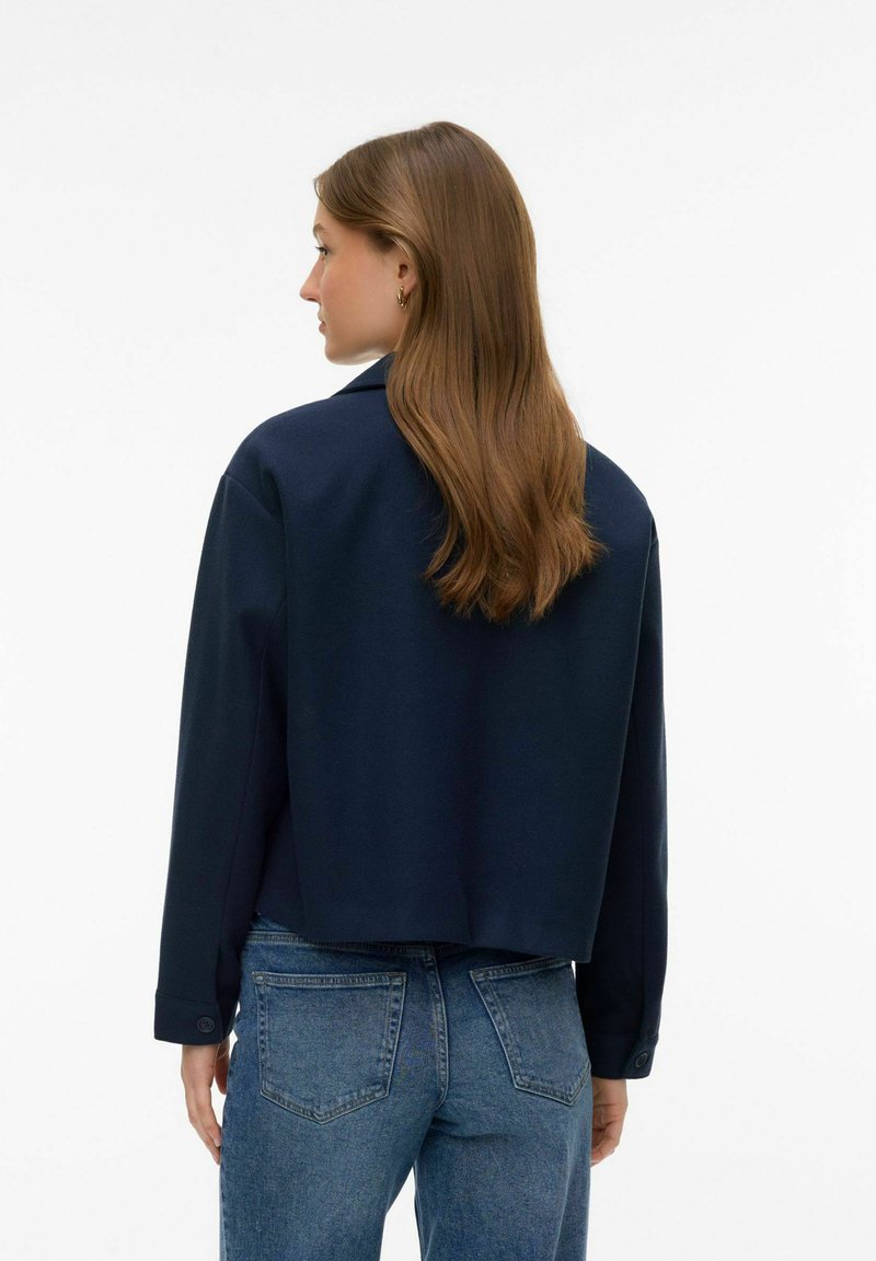 Femme aux longs cheveux bruns portant une veste bleu marine et un jean bleu, debout de dos contre un fond blanc.