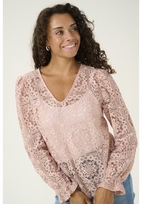 Blusa in pizzo rosa chiaro con scollo a V, caratterizzata da maniche a sbuffo e motivi geometrici. Texture morbida e vestibilità rilassata.