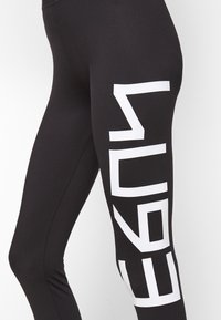 Svarta leggings i stretchigt tyg med djärv vit text längs sidan, vilket ger en slank och modern design.