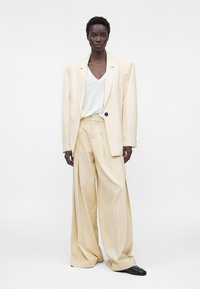 Mannequin aux cheveux courts portant un blazer crème oversize, un t-shirt blanc à col en V, un pantalon crème à jambes larges et des chaussures noires, debout devant un fond blanc.