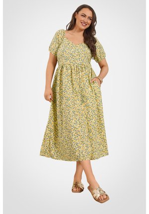 Frau lächelt, trägt ein gelbes Midi-Kleid mit grünem Blatt- und Blumenprint, kurzen Puffärmeln und goldenen Sandalen, posiert mit der Hand in der Tasche.