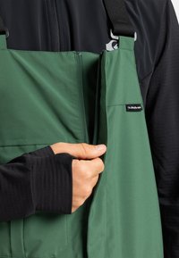 Een hand trekt de rits omhoog van een groene bretels, gedragen over een zwart langmouwhemd met duimopen in de manchetten.