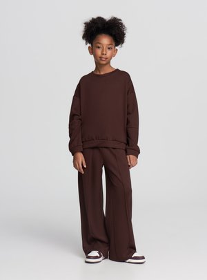 Felpa marrone con maniche extra large e orlo a coste, abbinata a pantaloni marroni a gamba larga. Scarpe da ginnastica bianche e marroni. Sfondo uniforme.