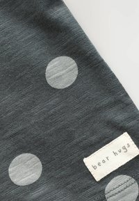Grå stof med en blød tekstur prydes af store lysegrå prikker. En label med teksten "bjørnekram" er fastgjort i nederste højre hjørne.