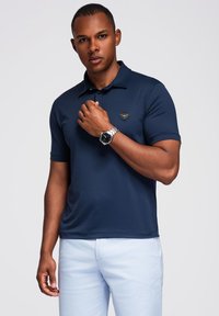 Uomo che indossa una polo blu navy e pantaloni chiari, mentre sistema un orologio argentato sul polso sinistro, in piedi davanti a uno sfondo neutro.