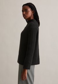 Schwarzer, gestrickter Pullover mit hohem Kragen, texturiertem Finish und langen Ärmeln, getragen über einer hellgrauen, gestreiften Hose.