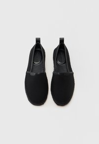 Zapatos negros sin cordones hechos de tela texturizada con acentos de cuero, que cuentan con una suela trenzada y lengüetas para poner y quitar fácilmente.