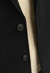 Abrigo de lana negro con un suéter texturizado beige debajo. Presenta un cuello con muesca y un detalle de botón en el lado izquierdo.