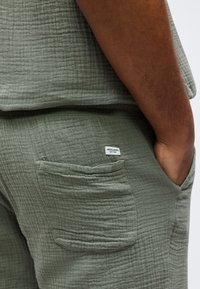 Κοντινό πλάνο χεριού ατόμου στην τσέπη υφασμάτινου, ελιάς-πράσινου παντελονιού με μια μικρή ορθογώνια ετικέτα Jack & Jones πάνω από την πίσω τσέπη.