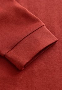 Langärmliges Textil mit geripptem Bund in einem einheitlichen Ziegelrot, mit einer weichen und glatten Textur. Keine sichtbaren Muster oder Beschläge.