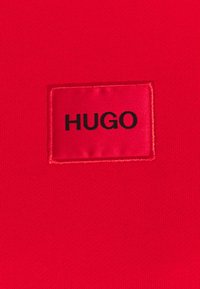 Textil rojo con una etiqueta rectangular cosida que lleva la palabra "HUGO" en letras negras negritas, rodeada por un fondo rojo a juego.