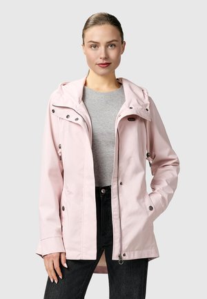 Kvinne iført en lys rosa hettejakke over en grå genser og svarte jeans, stående foran en ensfarget lys grå bakgrunn.