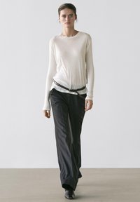 Kvinde går iført en gennemsigtig beige bluse med lange ærmer, mørke bukser med brede ben og tynde striber, sort bælte løst bundet og sorte sko.