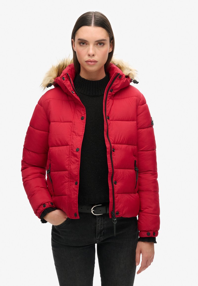 Donna che indossa un piumino rosso con cappuccio bordato di pelliccia, maglione nero e jeans scuri, in piedi con una mano in una tasca contro uno sfondo bianco.