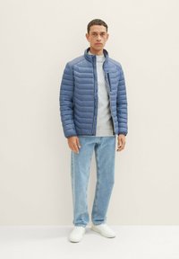 Pružená modrá bunda s vysokým golierom a zipsovým vreckom na hrudi, nosená cez sivú košeľu, skombinovaná s svetlomodrými džínsami a bielymi teniskami.