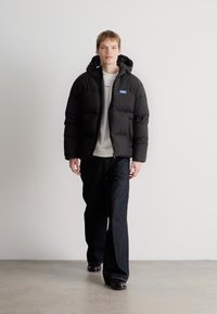 Μαύρο μπουφάν puffer με κουκούλα, διαθέτει μπροστινό φερμουάρ και δύο τσεπίδια. Φορεμένο πάνω από ένα λευκό πουκάμισο και σε συνδυασμό με σκούρα τζιν. Λείο υλικό.