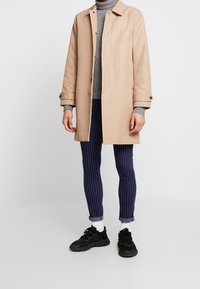 Beżowy płaszcz trencz z kołnierzem, z guzikami, na szarym swetrze. Do kompletu granatowe paskowane spodnie i czarne trampki z białymi skarpetkami.