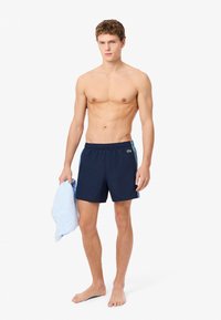 Mariene zwemshorts met elastische tailleband, zijzakken en blauwe accenten. Model houdt een lichtblauw handdoek vast, op blote voeten tegen een witte achtergrond.