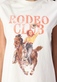 Krēmkrāsas topless t-krekls ar mirdzošu attēlu, kurā redzama sieviete, kas jāj uz brūnā zirdza, un virsū drukātas vārdi "RODEO CLUB" oranžā krāsā.