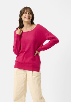 Fuchsia-Übergröße-Pullover mit weitem Ausschnitt, gerippten Bündchen und Saum. Weiche Textur mit subtilen Nähdetails. Kombination mit cremefarbenen Hosen.