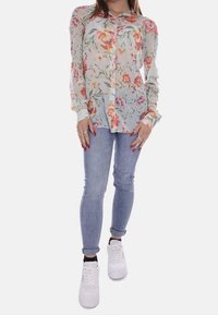 Blusa floreale a maniche lunghe in morbido tessuto trasparente con fiori multicolori; abbinata a jeans aderenti blu chiaro e sneakers bianche.