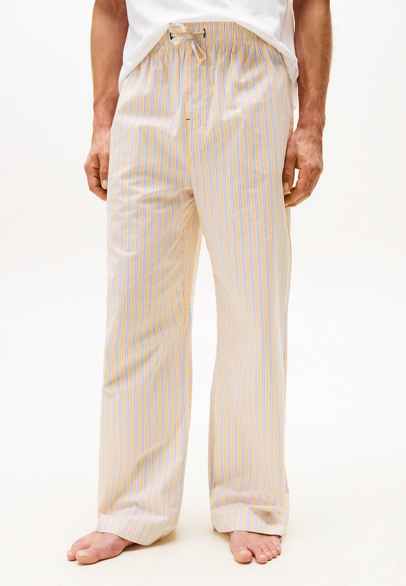 Man in losse beige en blauwe verticaal gestreepte pyjamabroek met een koord in de taille, staand op blote voeten tegen een effen achtergrond.