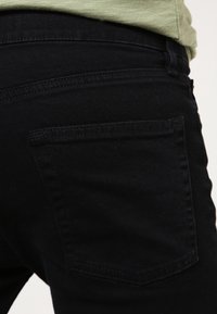 Jean noir avec une texture lisse, présentant une poche arrière et des détails de couture subtils le long de la taille et des bords de poche.
