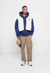 Fleece vest in cream over a blue hoodie, paired with beige cargo pants. Accessories include a tan cap and white sneakers.