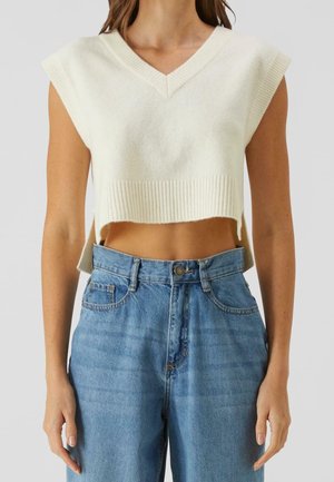 Femme portant un gilet court blanc sans manches en maille avec un col en V et un jean bleu taille haute sur un fond uni.