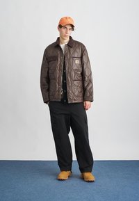 Chaqueta acolchada marrón con cuello oscuro y bolsillo, combinada con pantalones cargo negros y una gorra naranja, de pie sobre un fondo de alfombra azul.