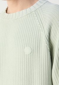 Gros plan d'un pull en maille texturée vert clair avec un col rond et un petit logo circulaire sur la poitrine.