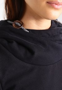 Närbild av nacke och nedre ansikte iförda en svart hoodie med en flätad snodd och ett runt metallspänne.