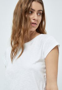 Camiseta blanca de manga corta con una tela suave y texturada y un escote redondeado. Presenta mangas enrolladas y sutiles arrugas para un detalle adicional.
