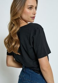 Jonge vrouw met lang golvend haar, een zwart T-shirt en blauwe spijkerbroek, kijkt over haar schouder tegen een effen achtergrond.