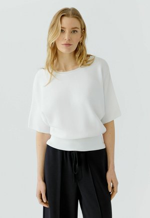 Femme aux longs cheveux blonds portant un pull blanc ample à manches courtes et un pantalon noir à cordon, debout devant un fond clair et uni.