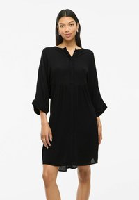 Robe chemise noire avec un col boutonné, manches longues retroussées aux poignets, silhouette décontractée et tissu léger.