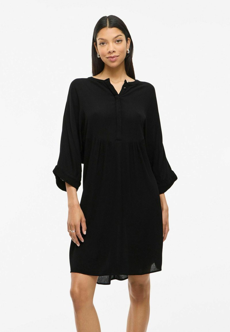 Robe chemise noire avec un col boutonné, manches longues retroussées aux poignets, silhouette décontractée et tissu léger.
