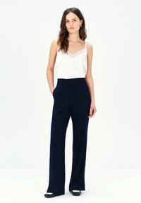 Weißes Spaghettitop mit Spitzenbesatz, kombiniert mit navyblauen, hochgeschnittenen, weit geschnittenen Hosen. Die Hosen verfügen über einen glatten Stoff und Seitentaschen.