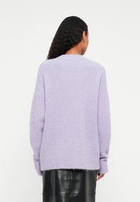 Tume lokkis juustega naine, kes kannab heledat lillakat karvast sviitrit ja musta nahks seelikut, vaadatuna tagant lihtsa tausta ees.