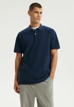 Młody mężczyzna w granatowej koszulce polo i jasnoszarych spodniach, stoi z jedną ręką w kieszeni na jednolitym białym tle.