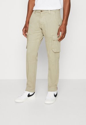 Beige cargo pants feature multiple utility pockets, straight-cut design, and stitched accents. Paired with white sneakers.