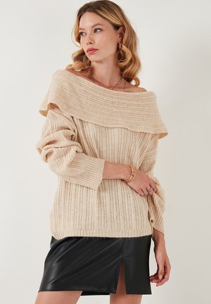 Maglione lavorato a maglia leggero color beige chiaro con scollo off-the-shoulder, texture a coste, maniche lunghe e vestibilità rilassata. Abbinato a una mini gonna nera in pelle.
