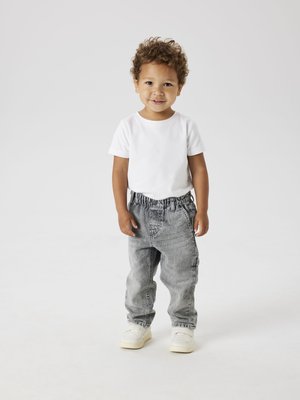 Kind in einem weißen Kurzarmhemd und grauen Jeanshosen mit elastischem Bund, stehend, mit hellfarbigen Turnschuhen.
