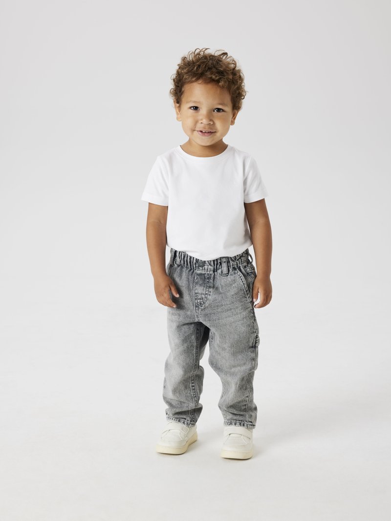 Niño con una camiseta blanca de manga corta y pantalones de mezclilla gris con cinturilla elástica, de pie, usando zapatillas ligeras.