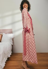 Ilga, rožinė raštuota chalatė su raudonais gėlių motyvais ir juostelėmis. Turi plačius rankovės ir atitinkamą juosmens raištį. Lygus audinys, iki kelių.