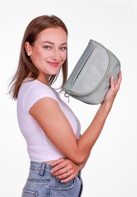 Šedá, zipová taška držená v ruce, s hladkým povrchem, zakulaceným tvarem a jemným logem na přední straně.