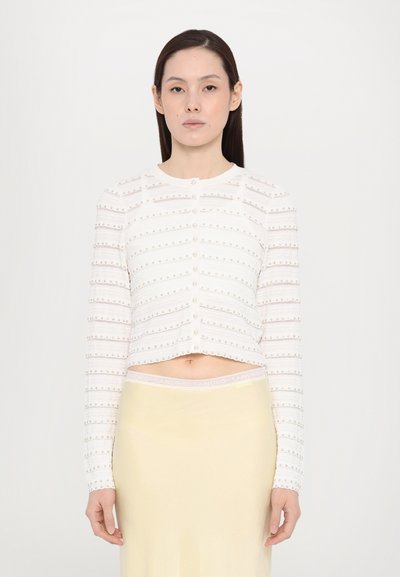 Bílý zkrácený cardigan s horizontálním pruhovaným vzorem a malými dekoračními knoflíky, spárovaný se světle žlutou sukní s logovaným pasem.