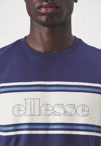 Navyblauwe T-shirt van katoen, voorzien van een grote witte streep met "ellesse" branding en twee horizontale lichtblauwe strepen.