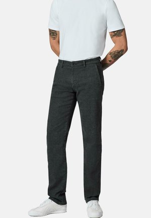 Männliches Model trägt dunkelgraue gerade geschnittene Hose, weiße Sneakers und ein weißes kurzärmeliges Hemd mit sichtbaren Tätowierungen an beiden Armen.
