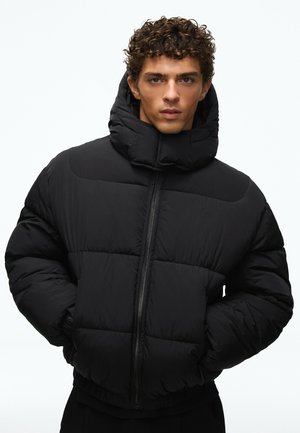 Jeune homme aux cheveux bouclés portant une doudoune noire à col montant et les mains dans les poches, sur un fond clair uni.